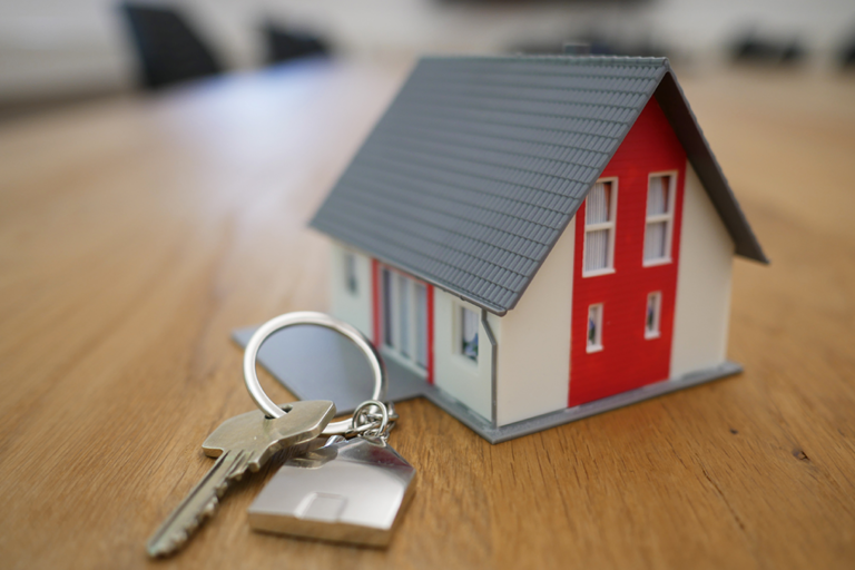 A photograph of a miniature model house with white and red paint and a gray shingle roof is dominant, with a set of keys on a keyring in the foreground.