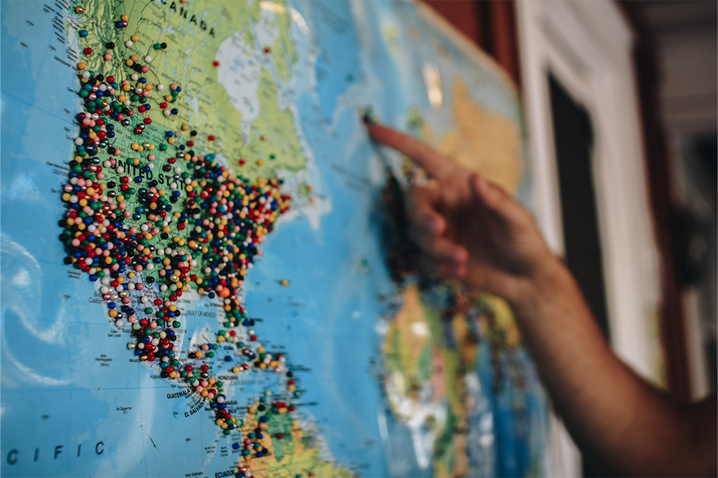 In the foreground is a a wall map of the United States with many pins covering it; in the background, a hand gesturing to other countries.