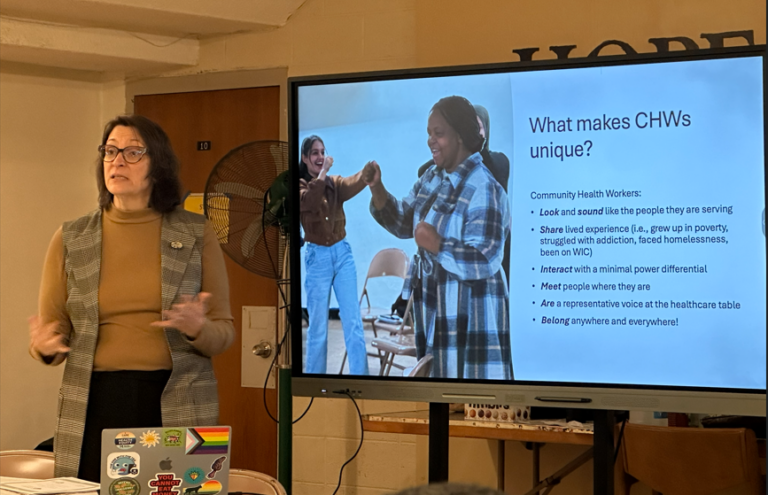 A woman in a plaid blazer presents next to a large screen. The slide, "What makes CHWs unique?" details Community Health Workers' roles. Her laptop has various stickers, adding a personal touch. The atmosphere is educational and engaging.