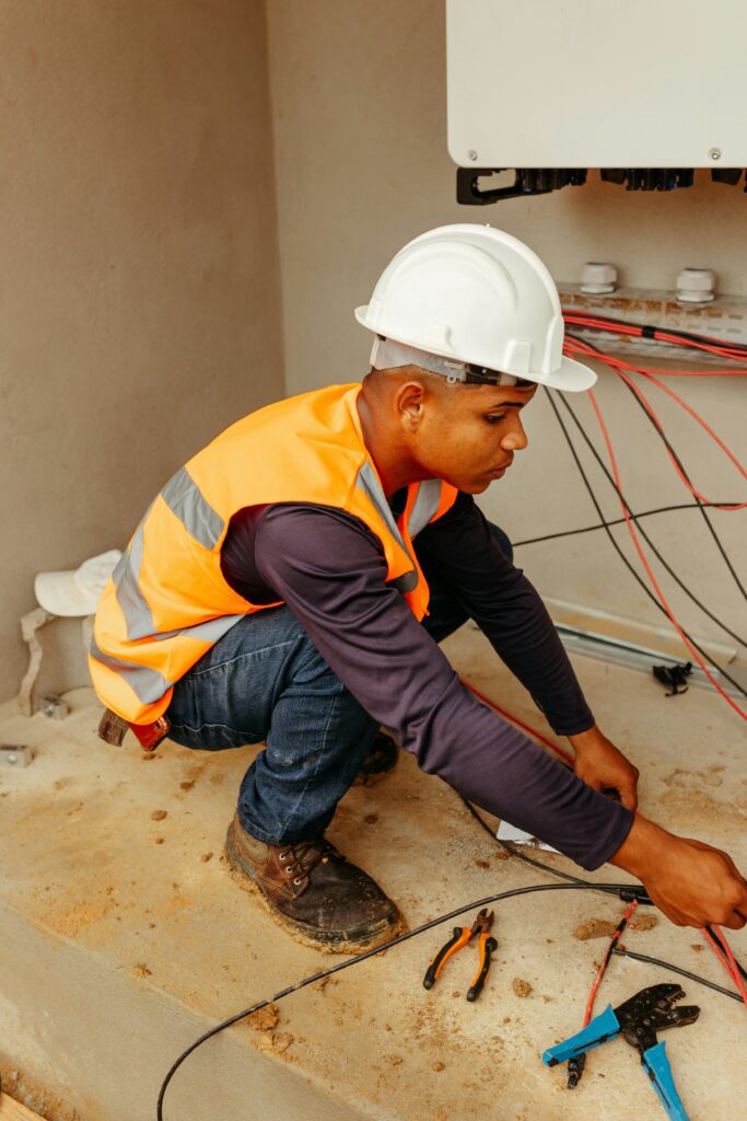 A construction worker wearing a white hard hat and orange safety vest squats, handling wires. Tools are scattered on the floor, conveying concentration.